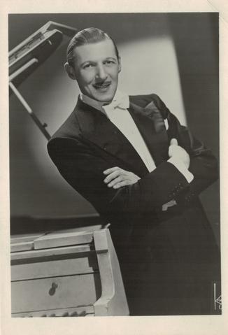 Caucasian male with mustache and slicked-back hair wearing a black tuxedo with white bowtie and leaning against a grand piano, ca. 1940s.